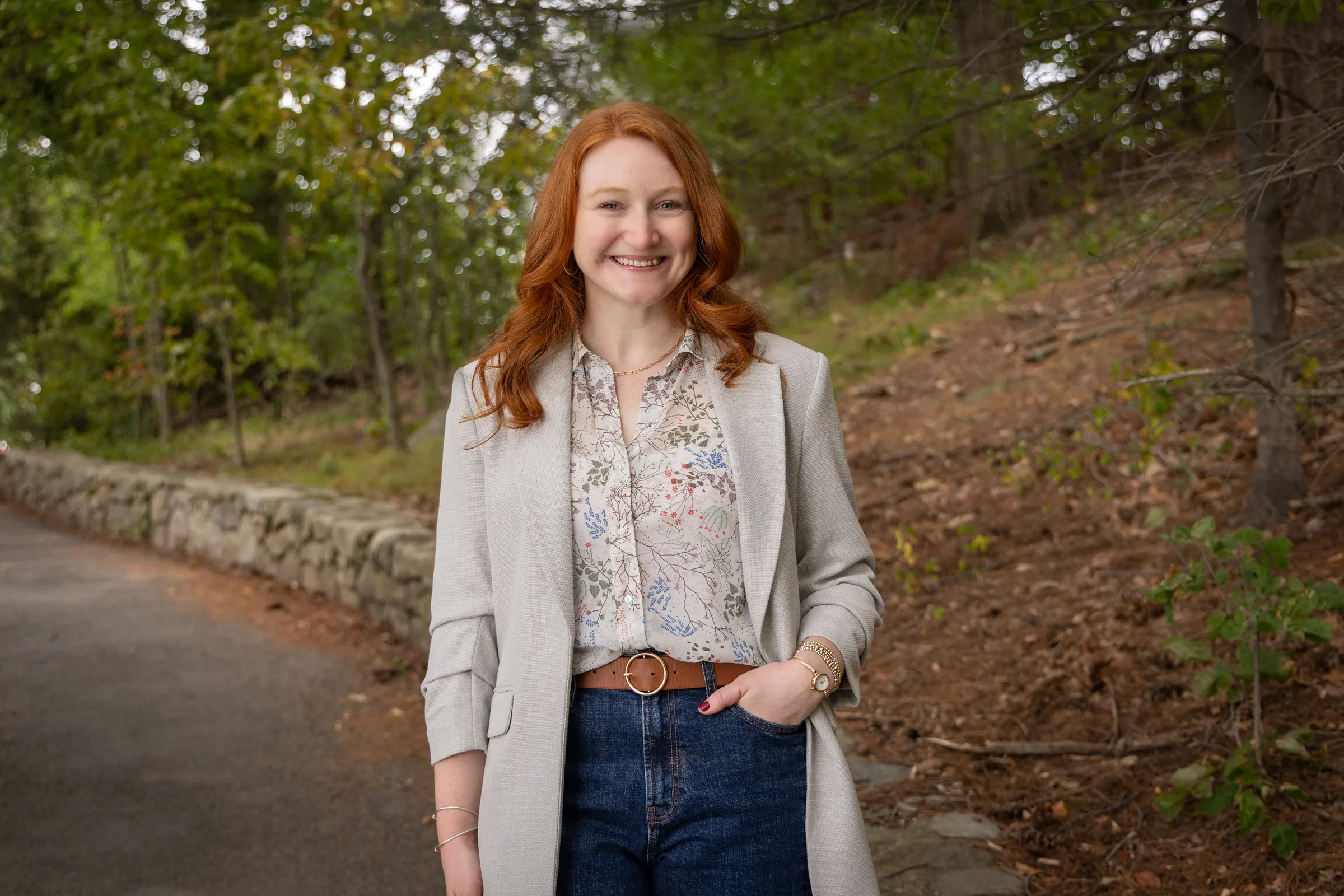 Professional headshot of a smiling woman with red hair in a light blazer on a wooded path